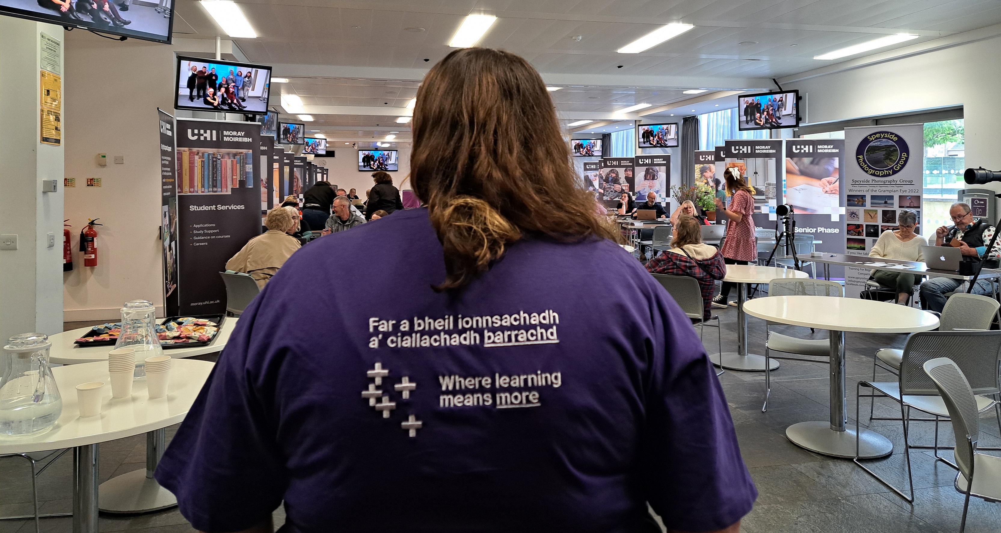 Photo of a person with their back faced to us looking into our AGBC Conference Room. The person is wearing a purple t-shirt which says Where Learning Means More in both English and Gaelic Photo of a person with their back faced to us looking into our AGBC Conference Room. The person is wearing a purple t-shirt which says Where Learning Means More in both English and Gaelic