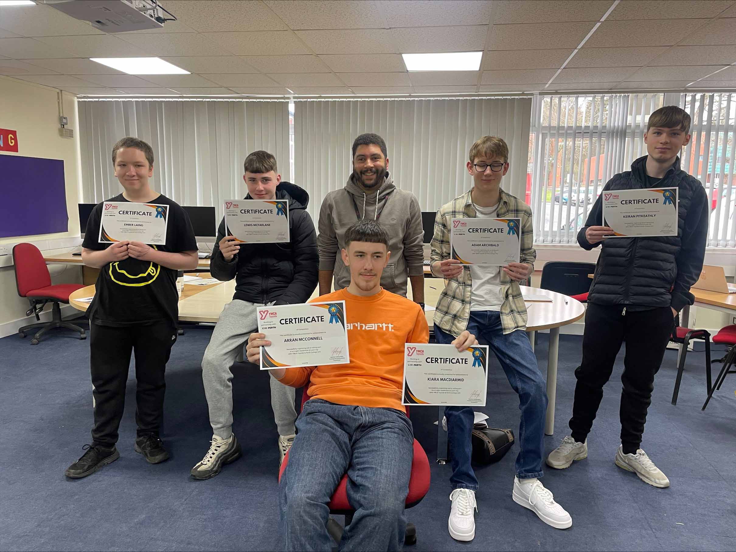 Six individuals are standing and sitting in a room, holding certificates. The room has tables, chairs, and a whiteboard in the background.