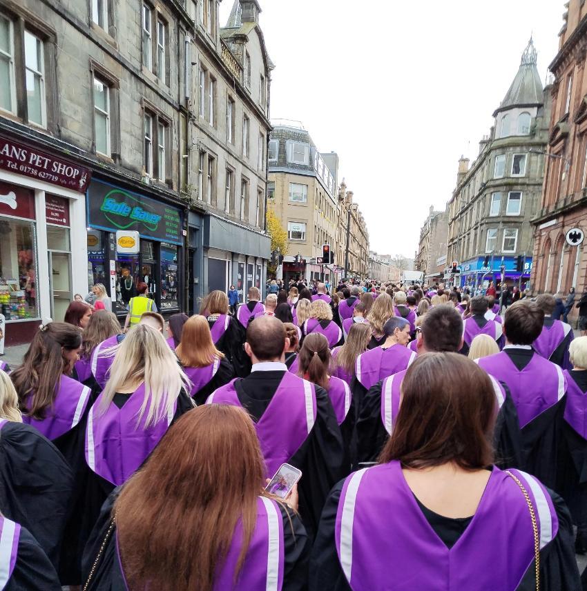 Group of UHI graduates clad in black gowns and purple hoods walking along a street with shop signs and shopfronts on show Group of UHI graduates clad in black gowns and purple hoods walking along a street with shop signs and shopfronts on show