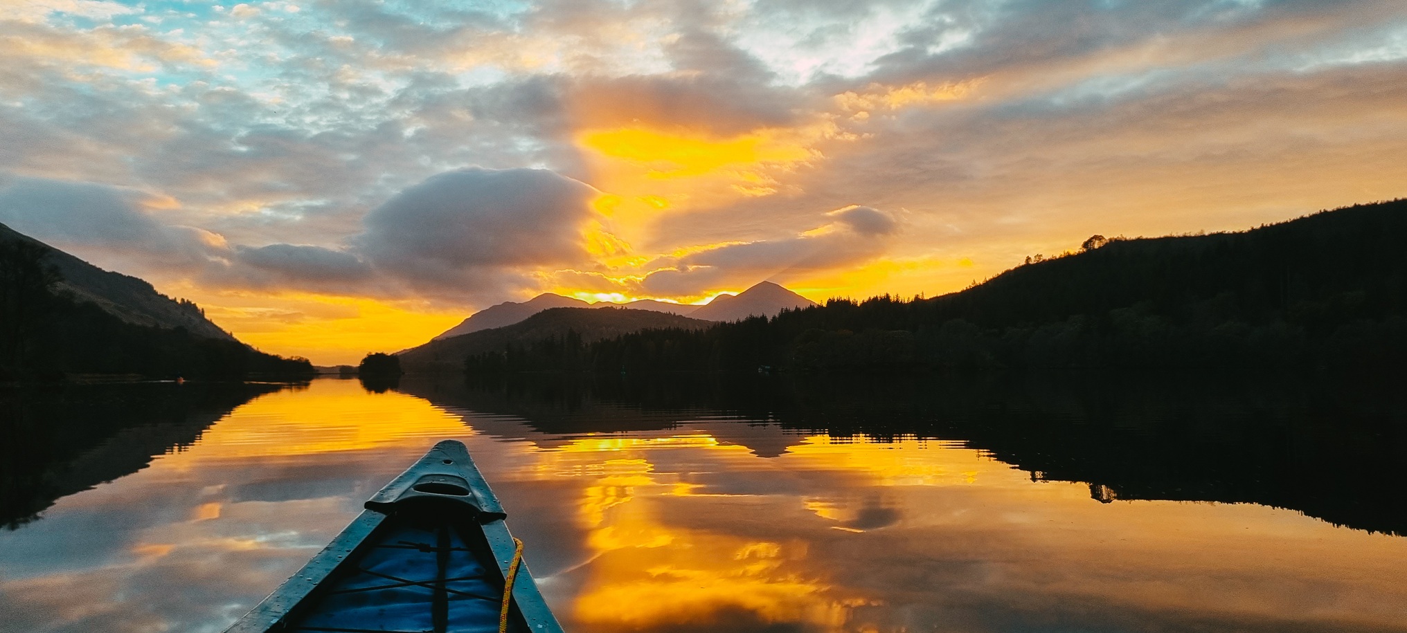 Photograph of a loch at sunset, with the dark outline of mountains in the distance against a sky with gold and orange clouds and glimpses of blue sky behind. In the foreground is the front part of a blue canoe, which is on the water. The sky and mountains are reflected in the still water.